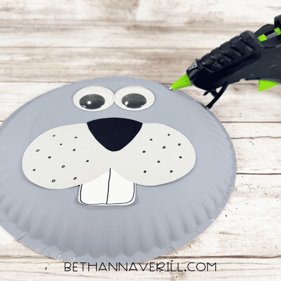Gray painted paper plate with googly eyes, a gray muzzle with black nose and dots, and a white tooth piece, with a hot glue gun nearby.