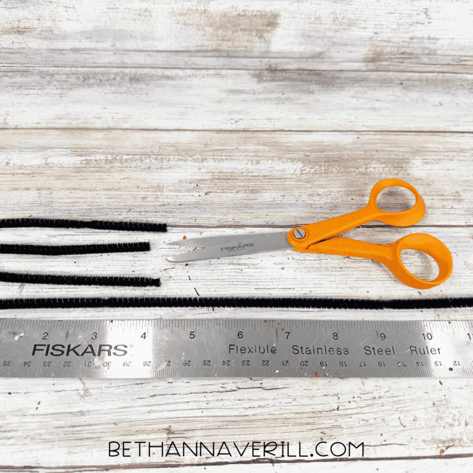 Black pipe cleaners cut into several shorter lengths beside orange scissors and a metal ruler.