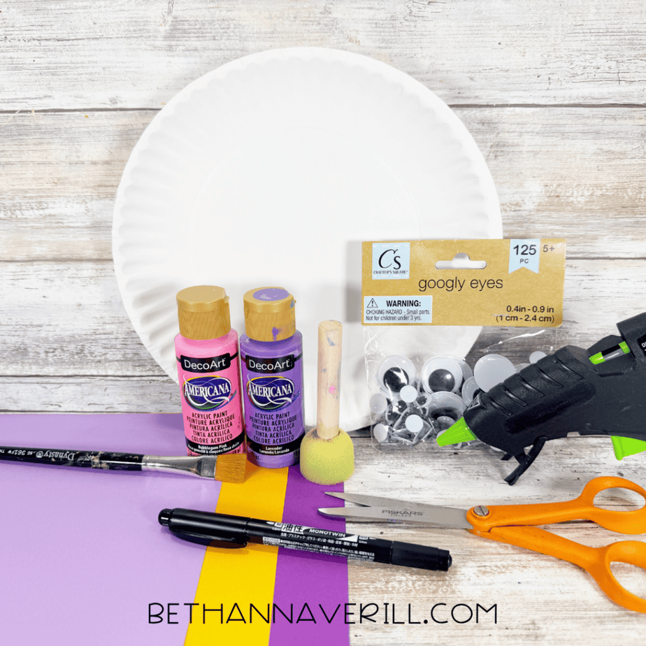 White paper plate with pink and purple acrylic paint, foam dauber, paintbrush, googly eyes, hot glue gun, orange scissors, black marker, and sheets of purple, lavender, and yellow paper.