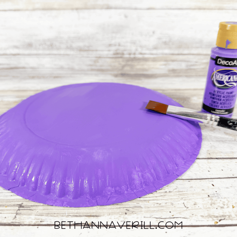 Paintbrush applying purple acrylic paint to the back of a paper plate, with a purple paint bottle beside it.