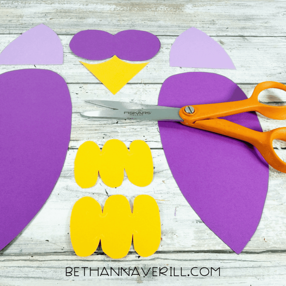 Cut paper pieces arranged on a white wood surface, including large purple wing shapes, lavender pointed pieces, a purple double-circle shape, yellow foot shapes, and a yellow pointed piece, with orange scissors nearby.