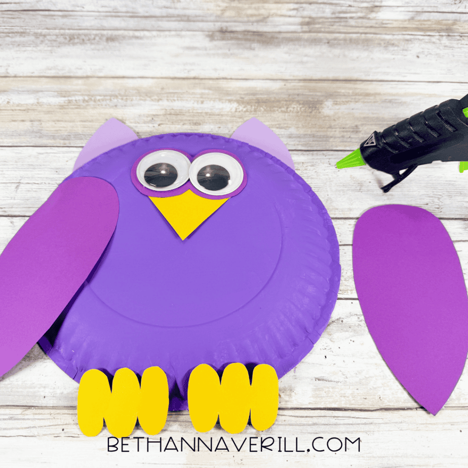 Partially assembled purple paper plate with lavender pointed pieces at the top, large googly eyes, a yellow pointed piece, purple wing pieces, and yellow foot pieces, with a hot glue gun nearby.