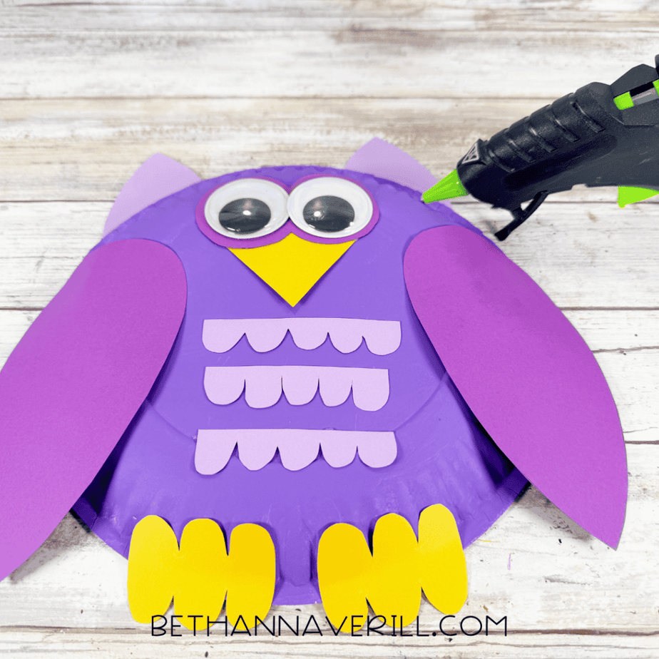 Purple paper plate with large purple wings, googly eyes, yellow feet, yellow pointed piece, and three rows of lavender scalloped paper pieces across the center.