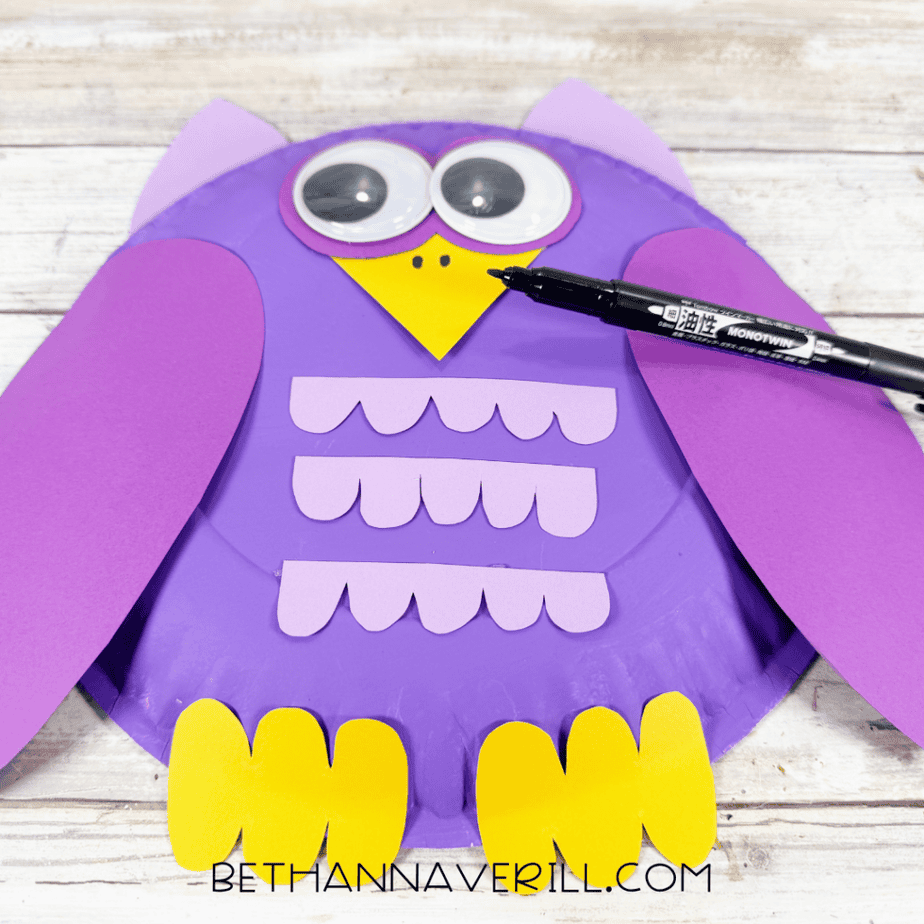 Purple paper plate owl with googly eyes, purple wings, lavender paper details, yellow feet and pointed beak, with two small black marks being added to the yellow paper with a black marker.