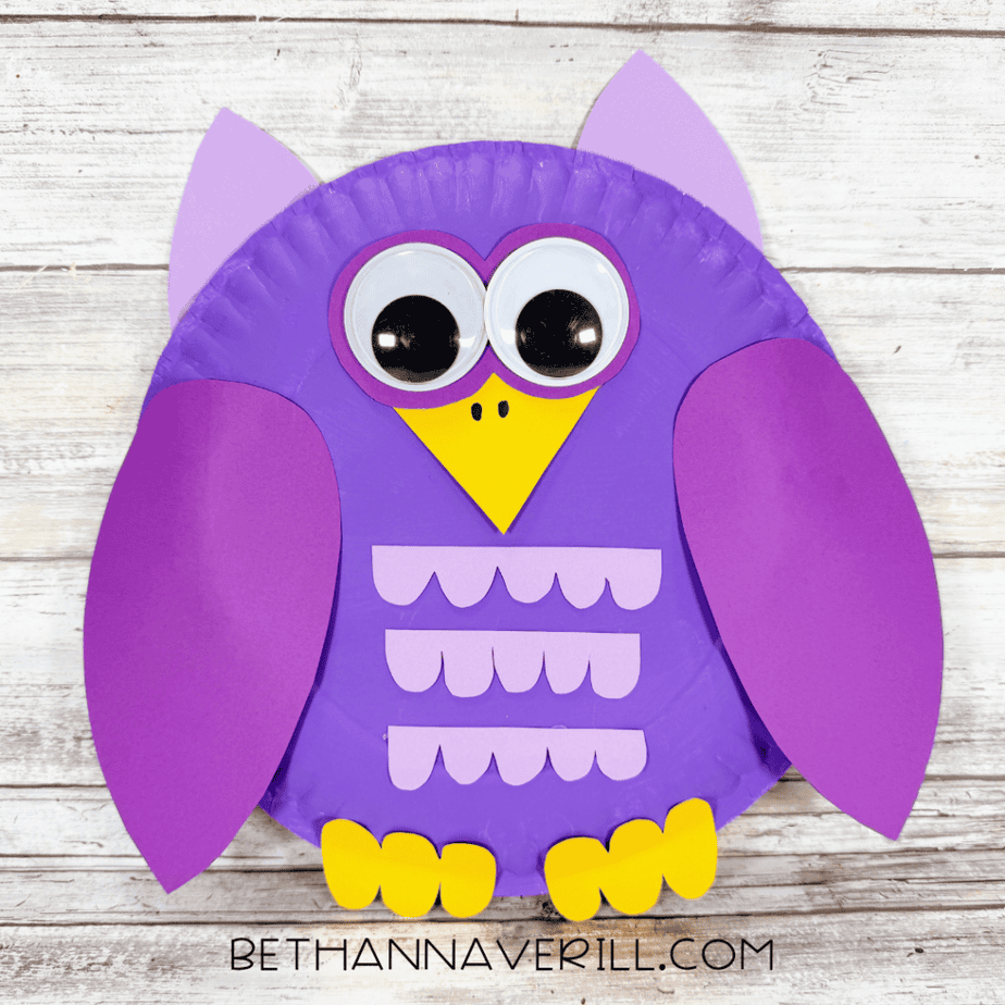 Finished paper plate owl with a purple body, large googly eyes, lavender pointed ears, purple wings, yellow beak and feet, and three rows of lavender scalloped paper pieces.
