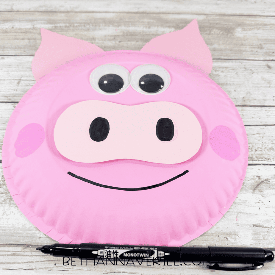 Pink paper plate face with googly eyes, black oval nostrils, pink cheeks, and a curved black smile, with a black marker below.