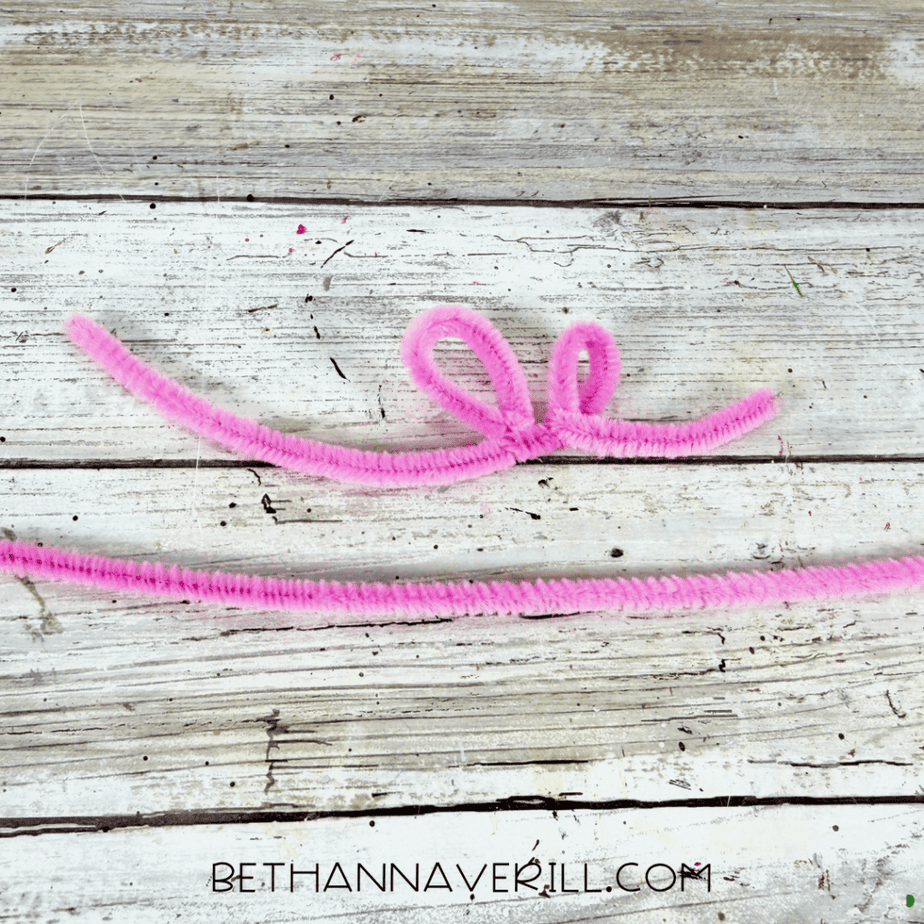 Pink pipe cleaner bent into a curly looped shape beside a straight pink pipe cleaner on a white wood surface.