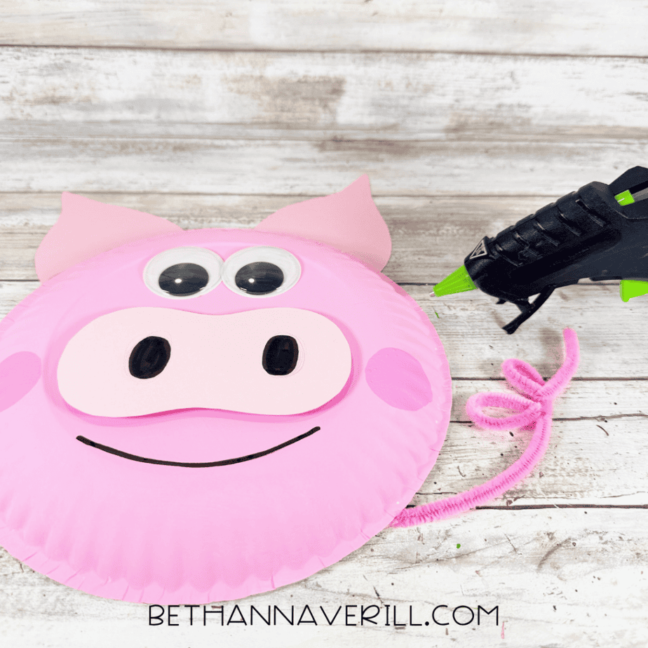 Pink paper plate pig face with googly eyes, pointed ears, light pink muzzle, black nostrils, pink cheeks, curved smile, and a curled pink pipe cleaner tail beside it.