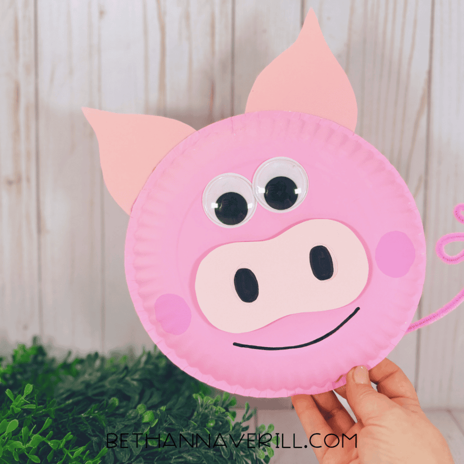 Finished pink paper plate pig craft held upright by a hand, with googly eyes, pointed ears, light pink muzzle, black nostrils, pink cheeks, smile, and curled pipe cleaner tail.
