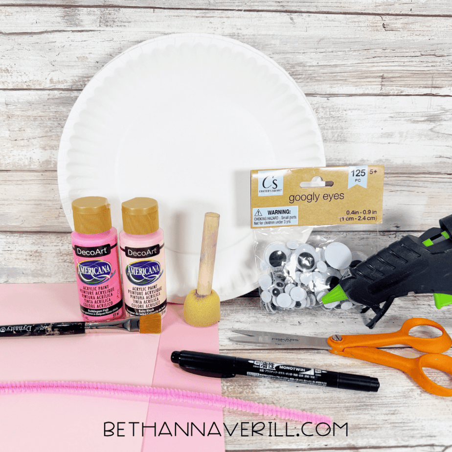 White paper plate with pink acrylic paint, paintbrush, foam dauber, googly eyes, hot glue gun, orange scissors, black marker, pink pipe cleaner, and sheets of pink paper.