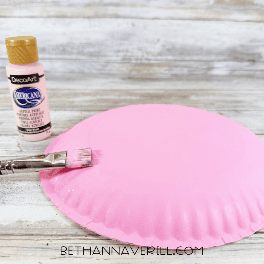 Paintbrush applying pink acrylic paint to the back of a paper plate, with a pink paint bottle beside it.