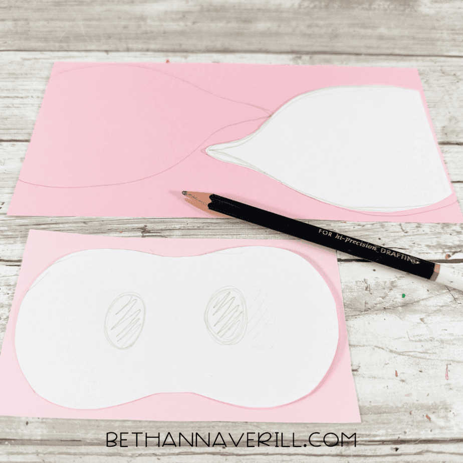 White paper templates traced onto pink paper, including two pointed ear shapes and a rounded muzzle shape, with a black pencil nearby.