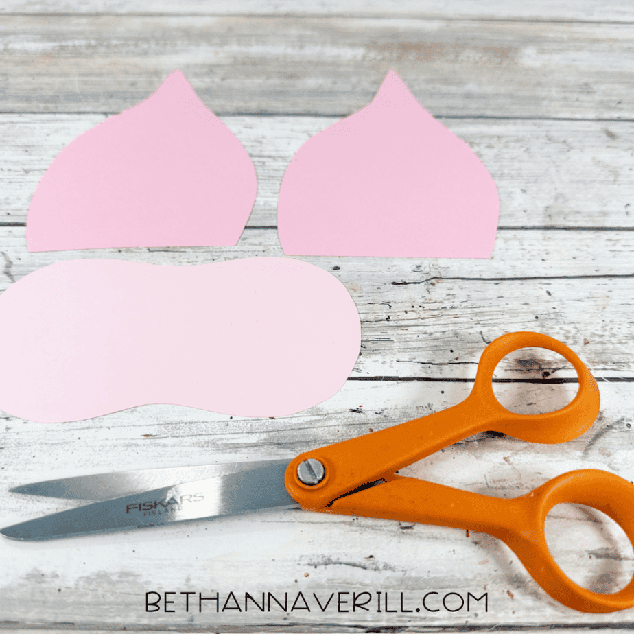 Two pink paper ear shapes and a rounded pink muzzle shape arranged beside orange scissors on a white wood surface.