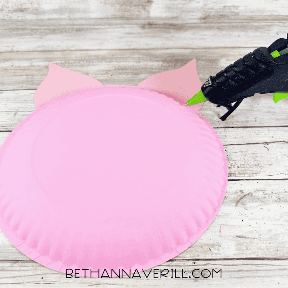 Pink painted paper plate with two pink paper ears attached at the top, with a hot glue gun nearby.