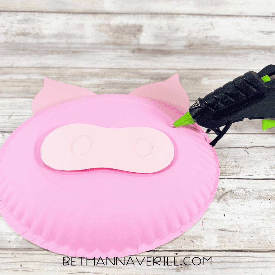 Pink painted paper plate with two ears and a lighter pink muzzle piece positioned on the front, with a hot glue gun nearby.