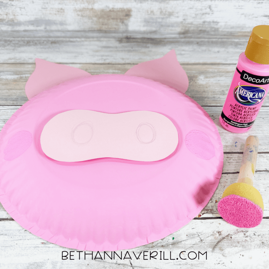 Pink paper plate face with a lighter pink muzzle, darker pink cheek circles, and a pink paint bottle and foam dauber beside it.