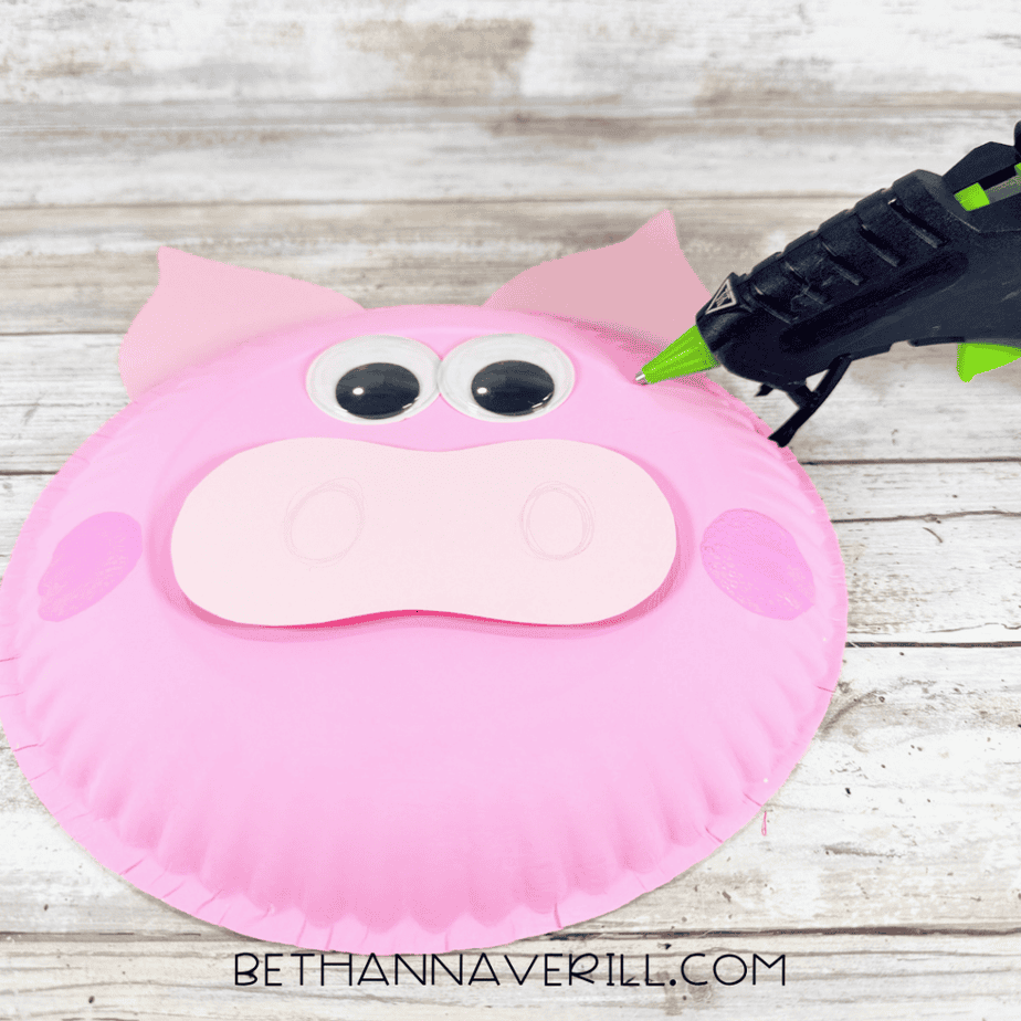 Pink paper plate face with two large googly eyes, a light pink muzzle, darker pink cheeks, and a hot glue gun nearby.