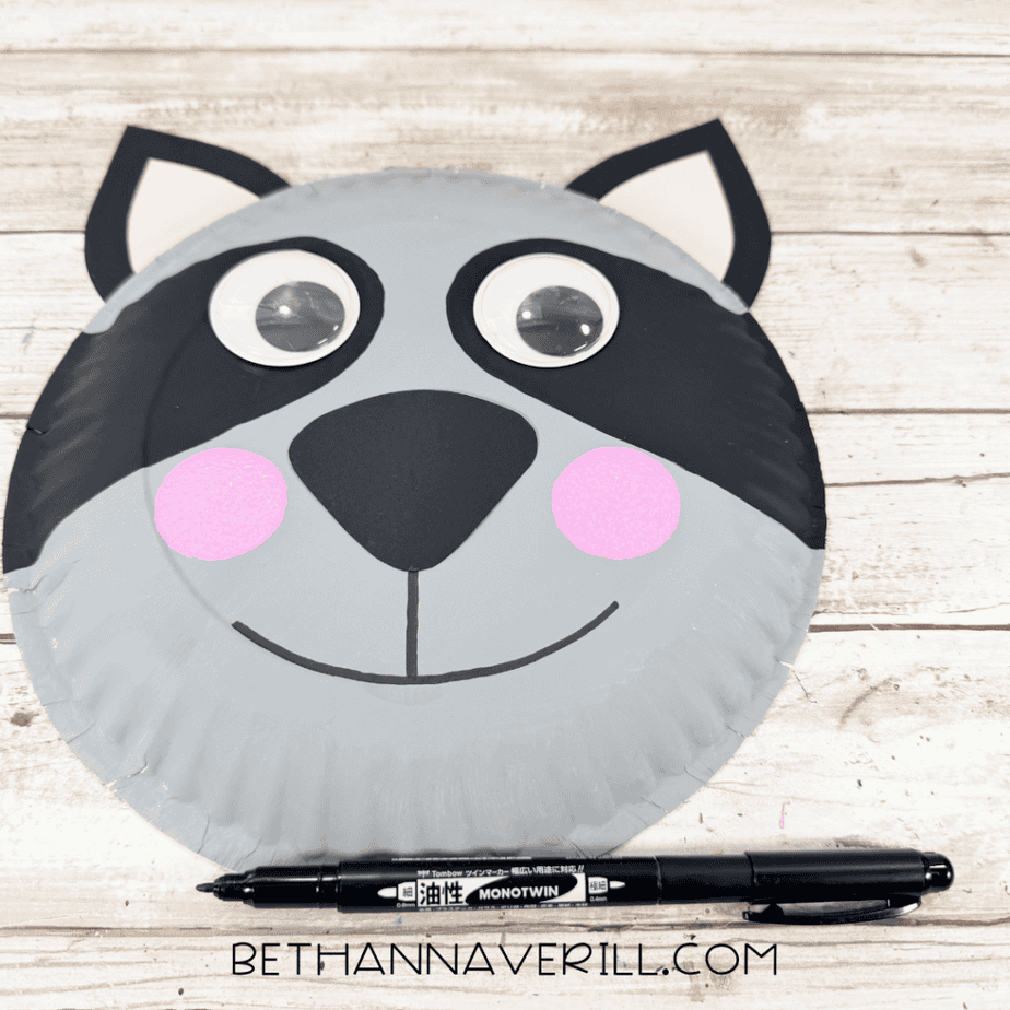 Gray paper plate face with black eye markings, googly eyes, pink cheeks, black nose, and a curved black mouth, with a black marker below.