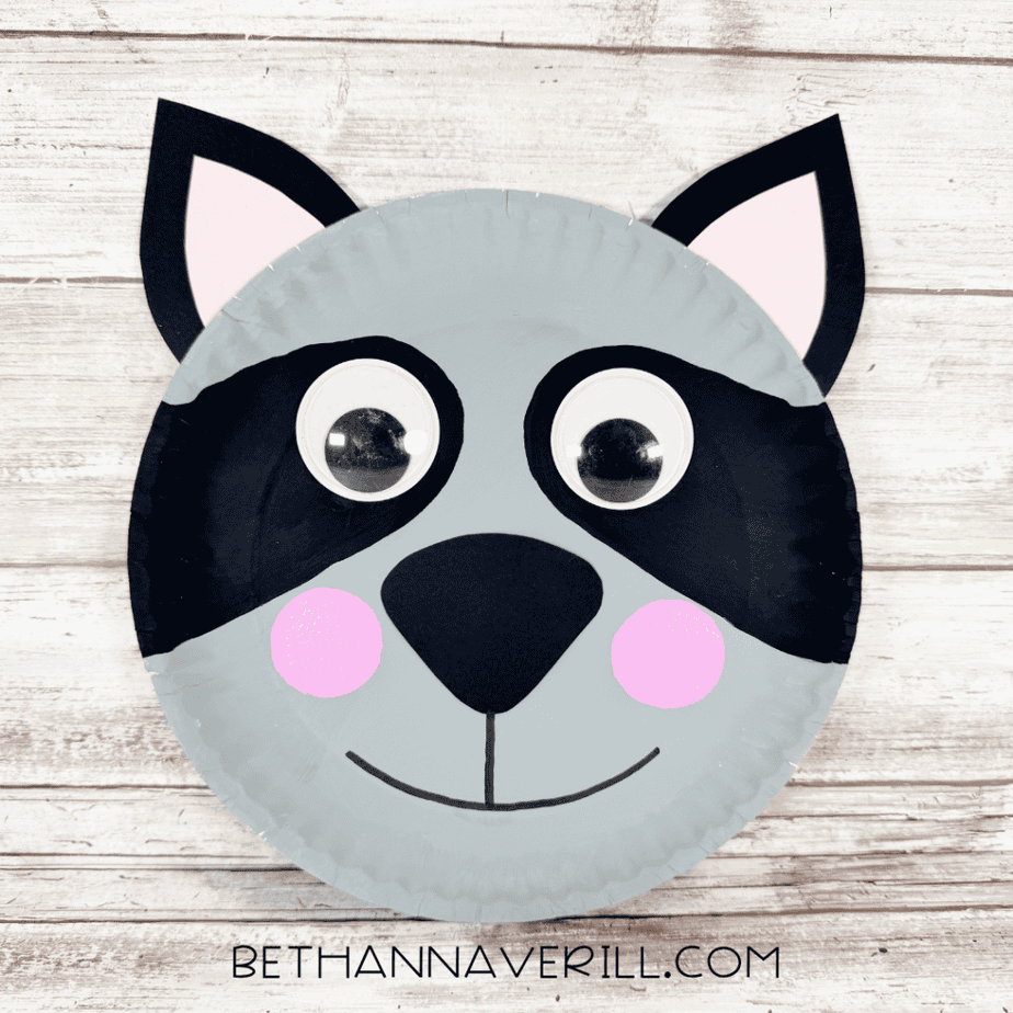 Finished paper plate raccoon face with gray coloring, black eye markings, pink-centered ears, large googly eyes, black nose, pink cheeks, and a smiling mouth.