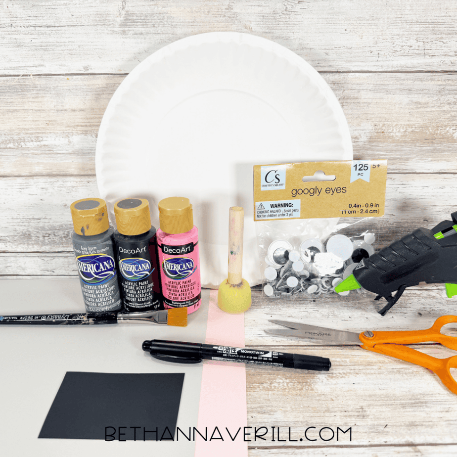 White paper plate with gray, black, and pink acrylic paint, googly eyes, foam dauber, paintbrush, hot glue gun, orange scissors, black marker, and sheets of gray, black, and pink paper.
