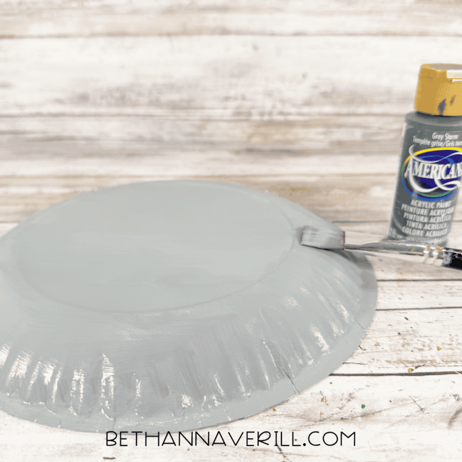 Paintbrush applying gray acrylic paint to the back of a paper plate, with a gray paint bottle beside it.