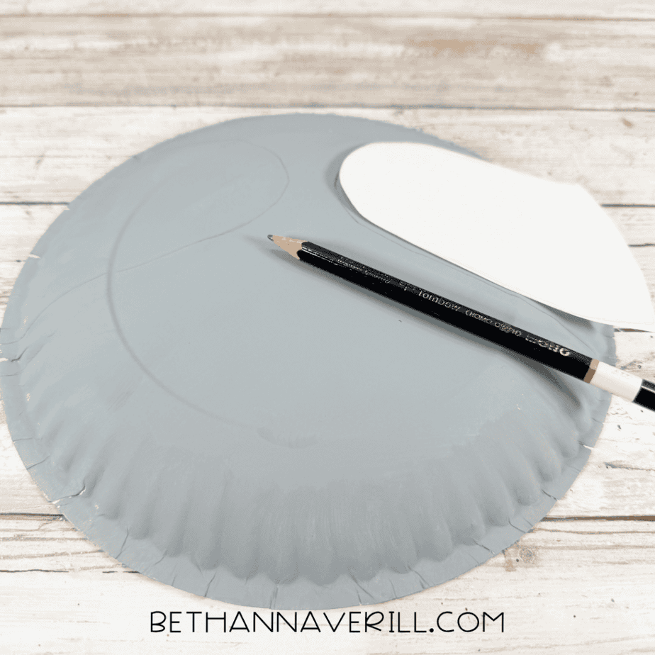 Gray painted paper plate with a white paper template positioned near the top, traced curved lines, and a black pencil across the plate.