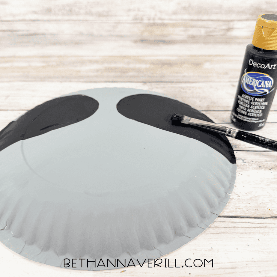 Black acrylic paint being brushed onto two curved sections of a gray paper plate, with a black paint bottle beside it.