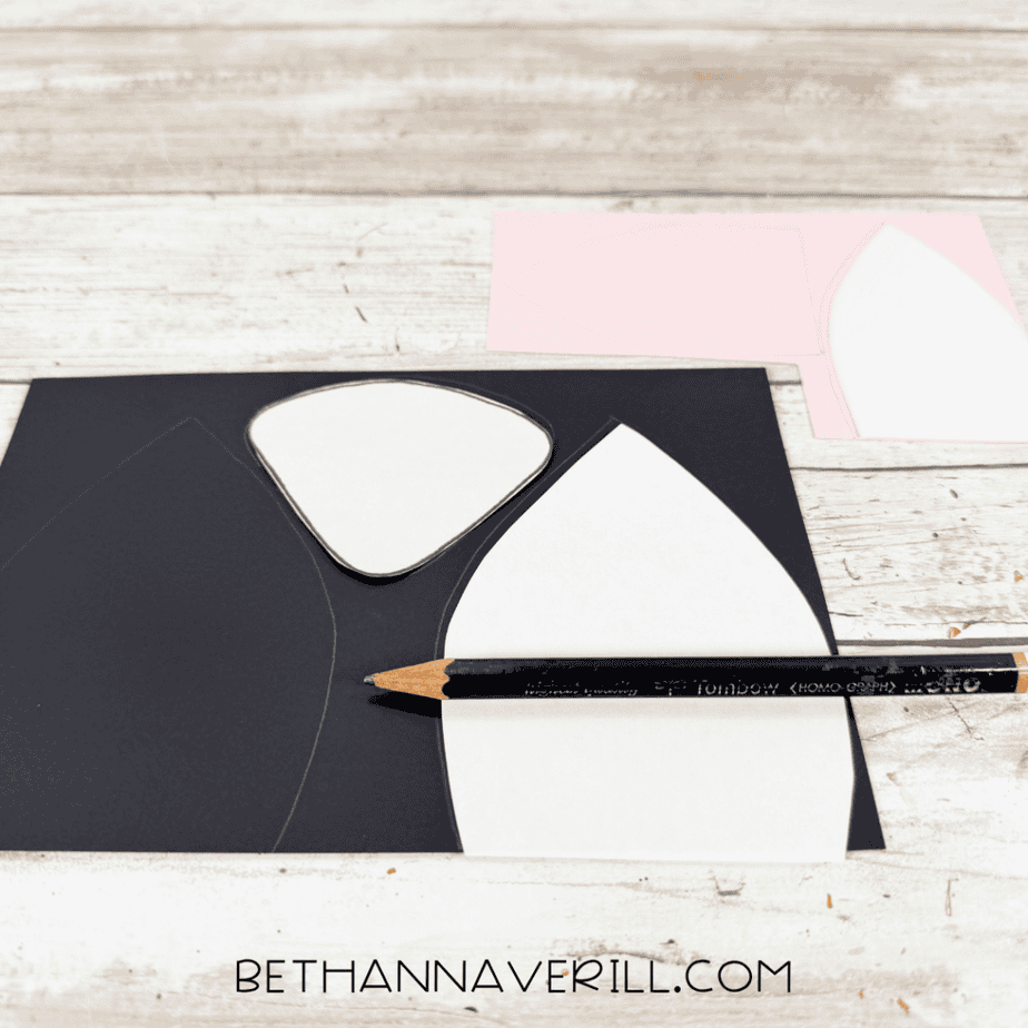 White paper templates traced onto black and pink paper, including pointed ear shapes and a rounded triangular shape, with a black pencil nearby.