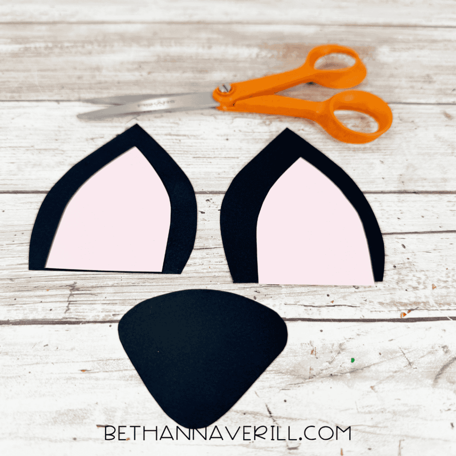 Two black paper ear shapes with pink centers and a rounded black paper nose shape arranged below orange scissors.