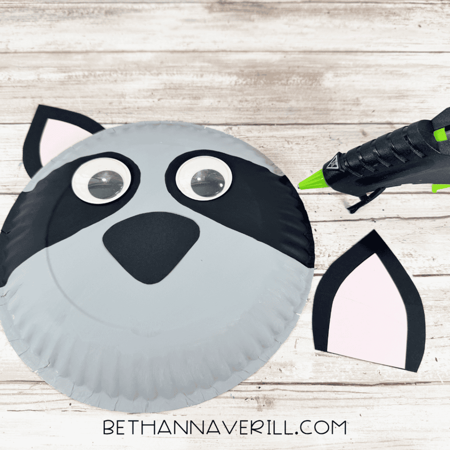Partially assembled gray paper plate face with black painted sections around large googly eyes, a black paper nose, and one attached pink-centered ear, with another ear and hot glue gun beside it.