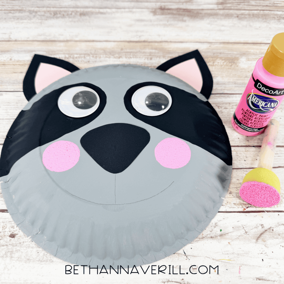 Gray paper plate face with two pink-centered ears, black eye markings, large googly eyes, black nose, and two pink painted cheek circles, with pink paint and a foam dauber beside it.
