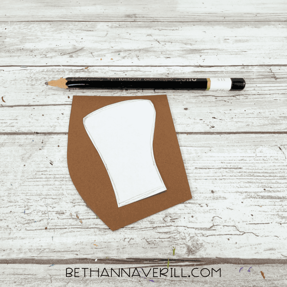 White paper template of an acorn stem placed on brown construction paper with a pencil above it.