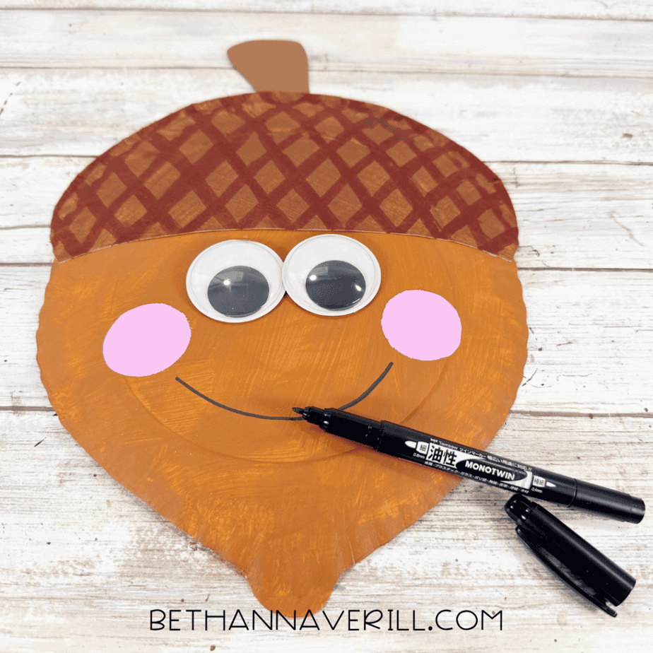Black marker drawing a curved smile onto an orange acorn paper plate craft with googly eyes and pink cheeks.