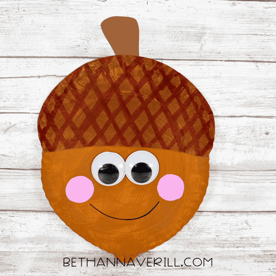 Finished smiling acorn paper plate craft featuring a patterned brown cap, large googly eyes, pink cheeks, and a brown paper stem.