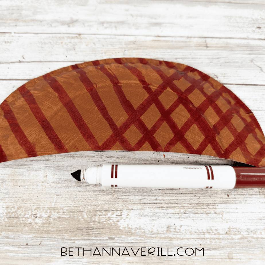 Brown painted paper plate semicircle decorated with a crisscross pattern using a dark brown marker.