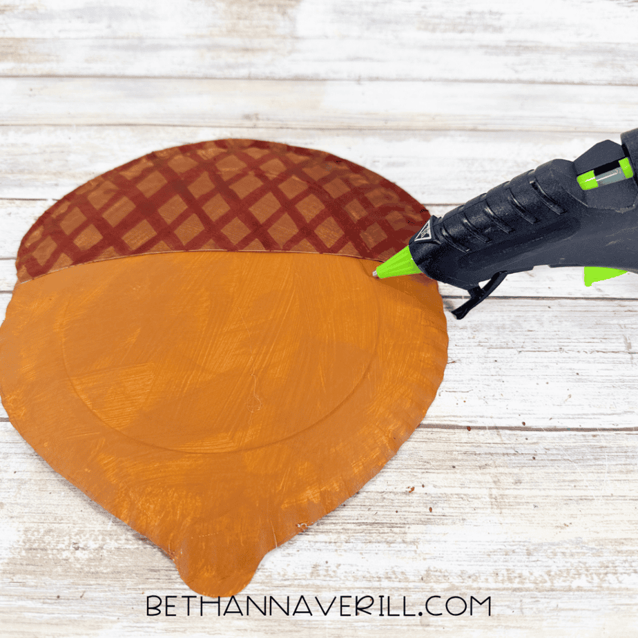 Hot glue gun attaching a patterned brown paper plate piece to the top of an orange acorn-shaped paper plate.