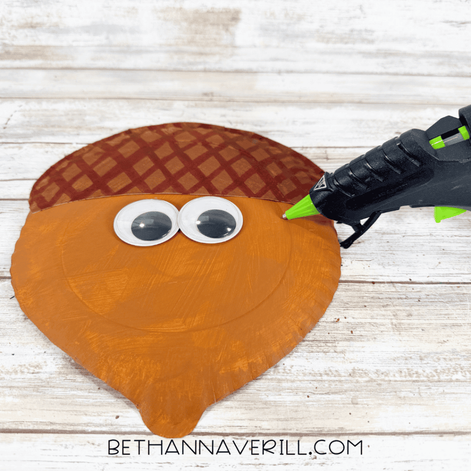 Two large googly eyes being glued onto an orange paper plate acorn beneath its patterned brown cap.
