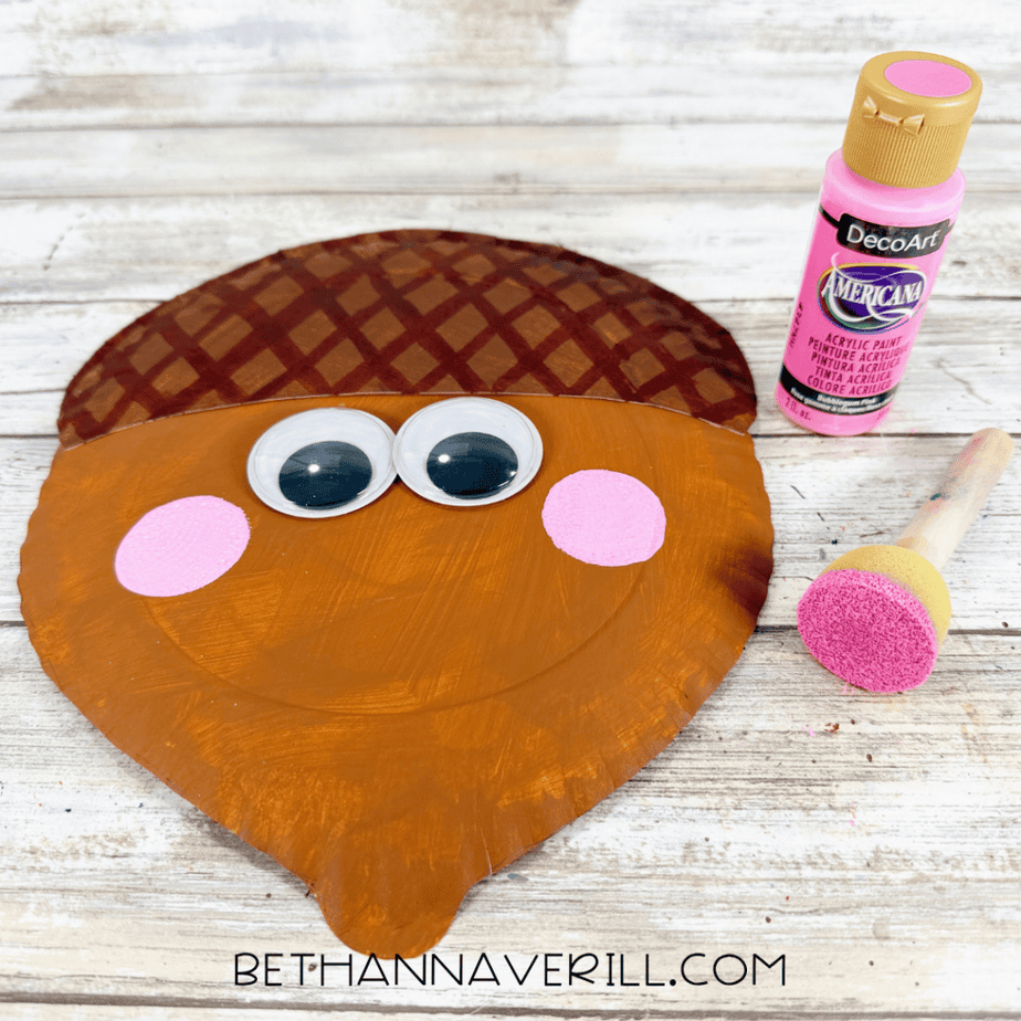Pink paint and a round sponge brush used to add rosy cheeks to a smiling acorn paper plate craft.