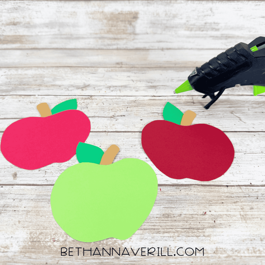 Three colorful paper apples assembled with green leaves and brown stems beside a hot glue gun.