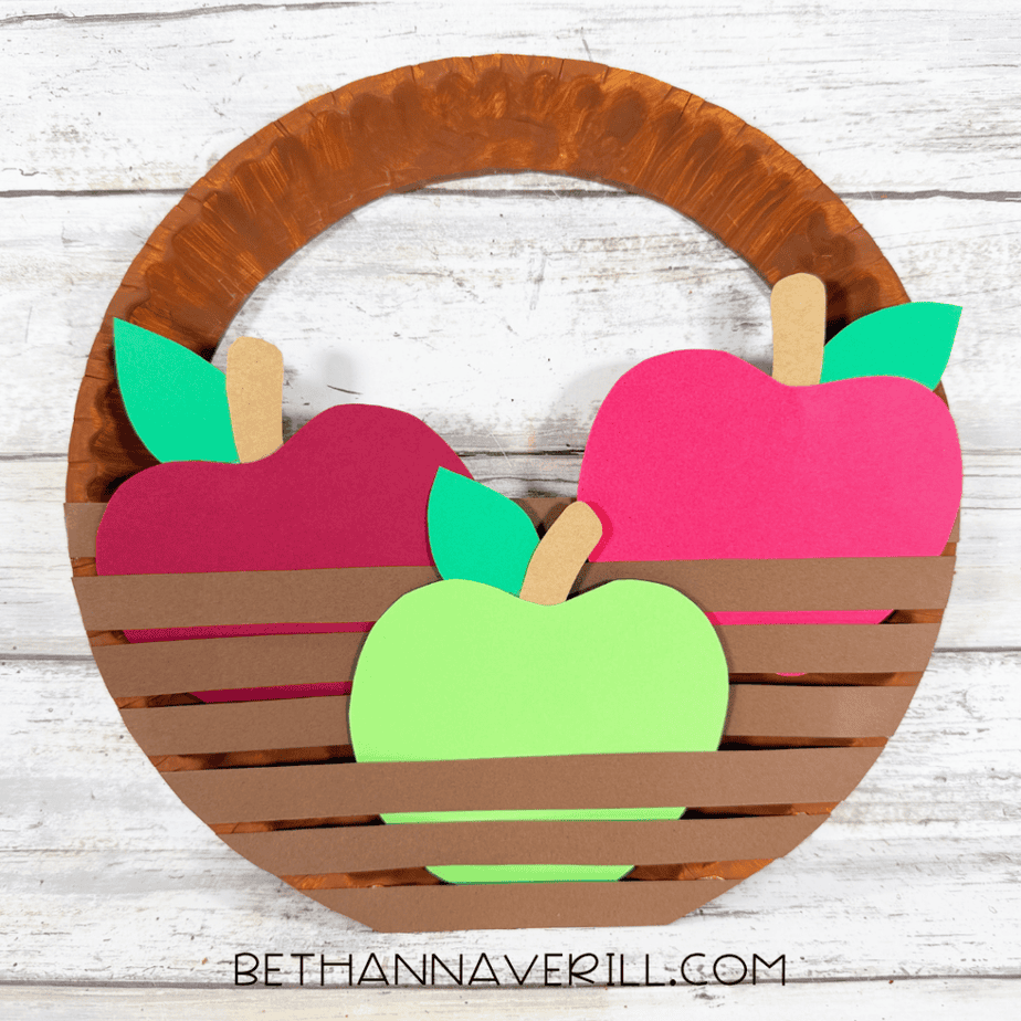 Finished paper plate apple basket craft filled with red, pink, and green construction paper apples behind brown paper strips.