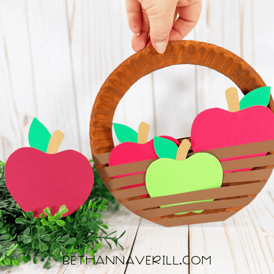 Hand holding a completed brown paper plate apple basket filled with colorful paper apples beside a red paper apple and greenery.