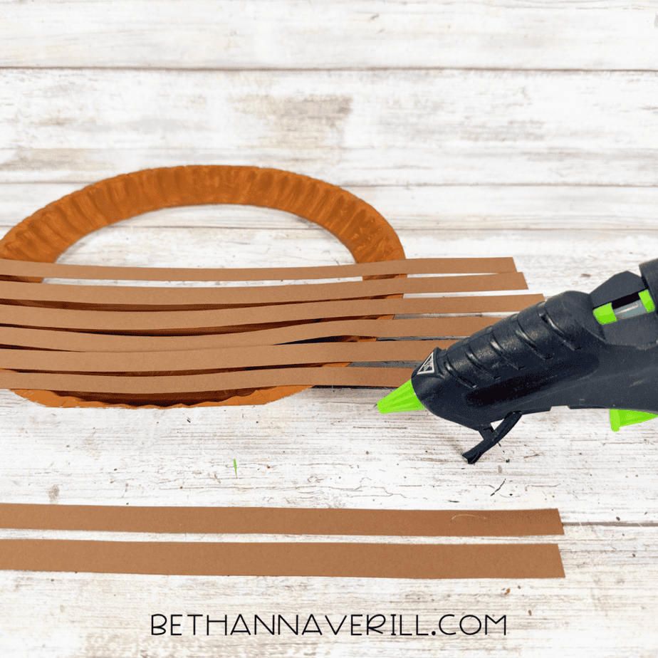 Brown construction paper strips arranged horizontally across a painted paper plate basket with a hot glue gun nearby.