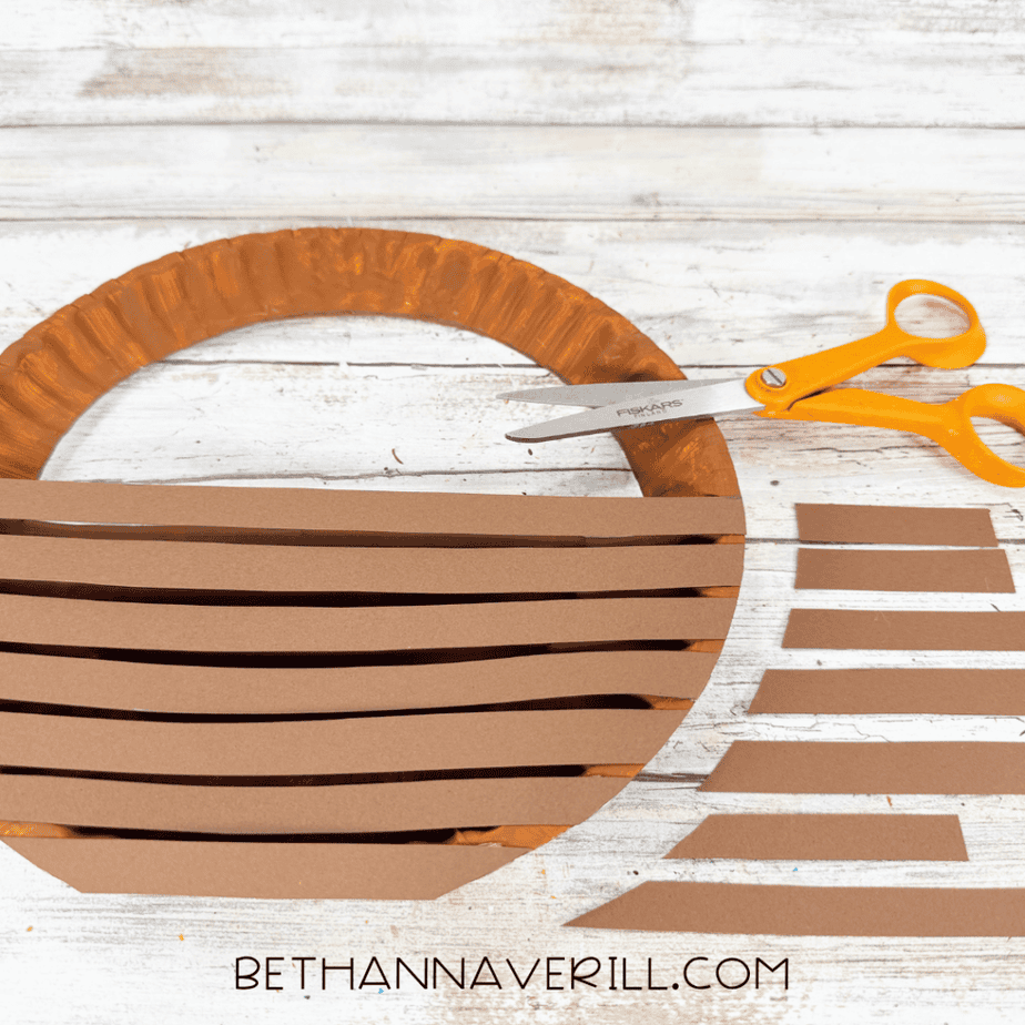 Brown paper strips trimmed to fit the rounded edges of a paper plate apple basket using scissors.