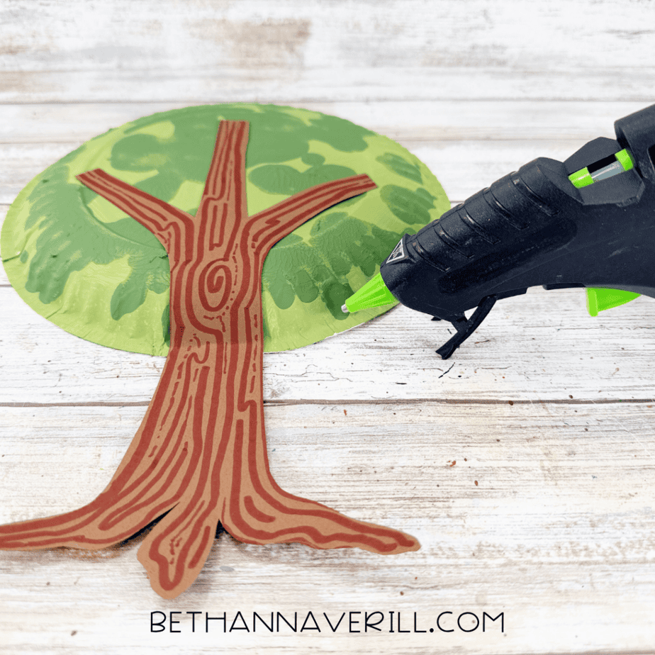Hot glue gun beside a brown construction paper tree trunk positioned over a green painted paper plate canopy.