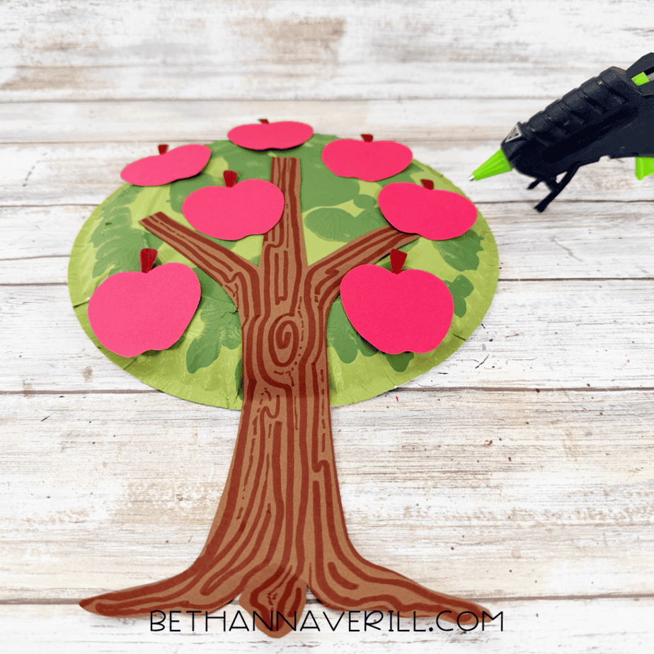 Red construction paper apples arranged across a green painted paper plate tree with a brown paper trunk and hot glue gun nearby.