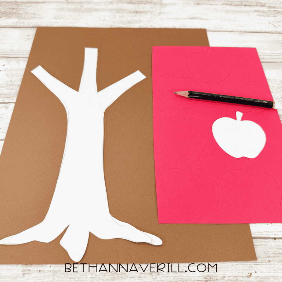 Paper tree trunk and apple templates placed on brown and red construction paper with traced outlines and a pencil.