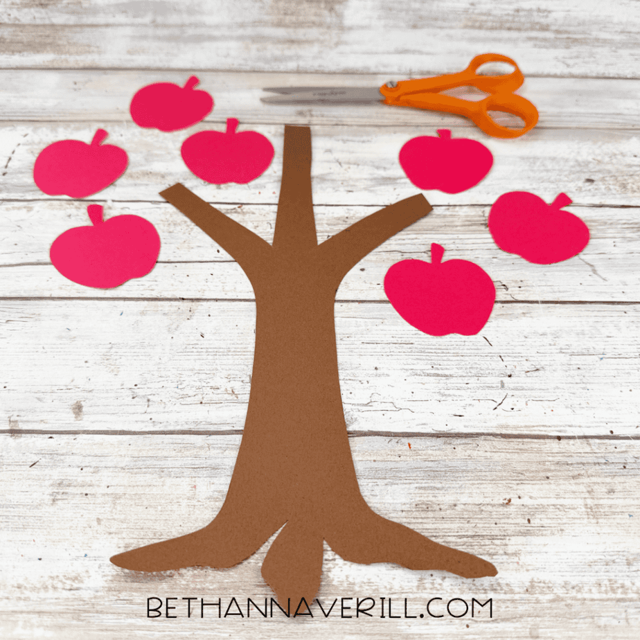 Brown construction paper tree trunk surrounded by six red paper apple cutouts and orange scissors.