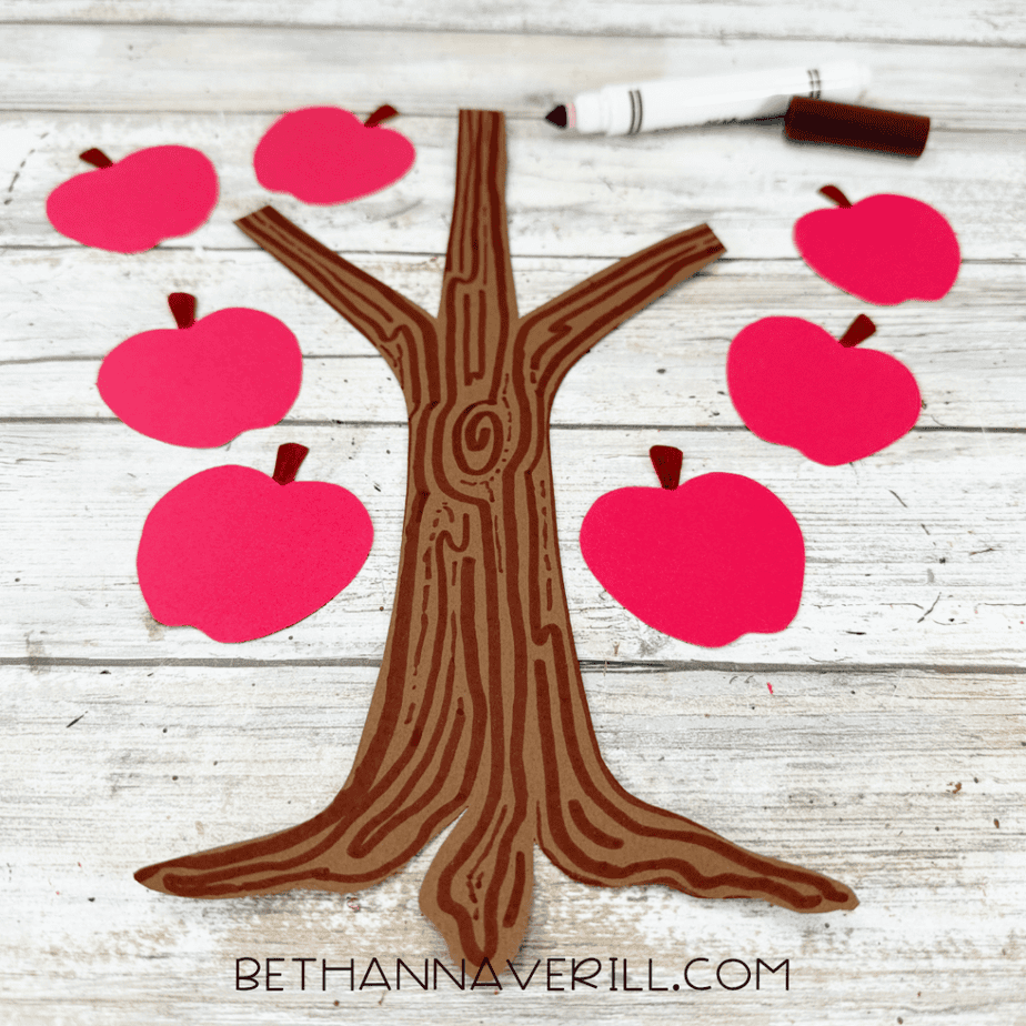 Brown paper tree trunk decorated with dark brown marker lines to create a wood grain pattern, surrounded by red paper apples.