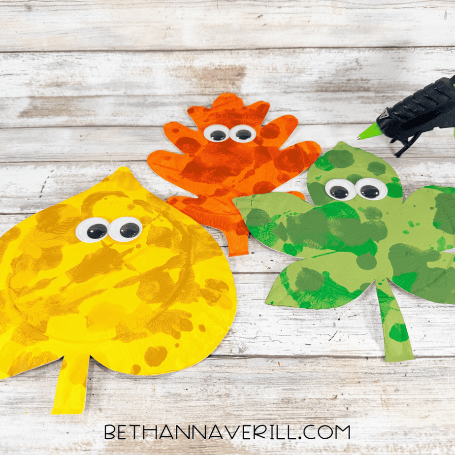 Orange, yellow, and green paper plate leaves decorated with large googly eyes, with a hot glue gun nearby.