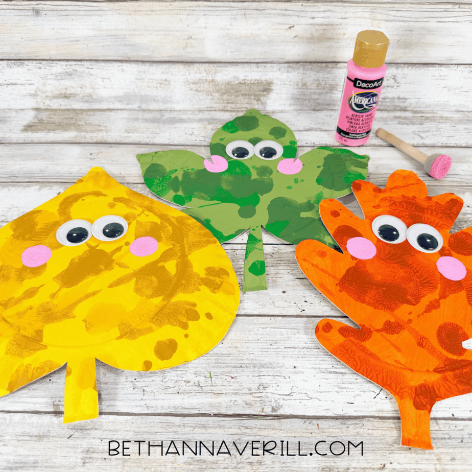 Three colorful paper plate autumn leaves with googly eyes and pink painted cheeks beside a pink paint bottle and sponge brush.